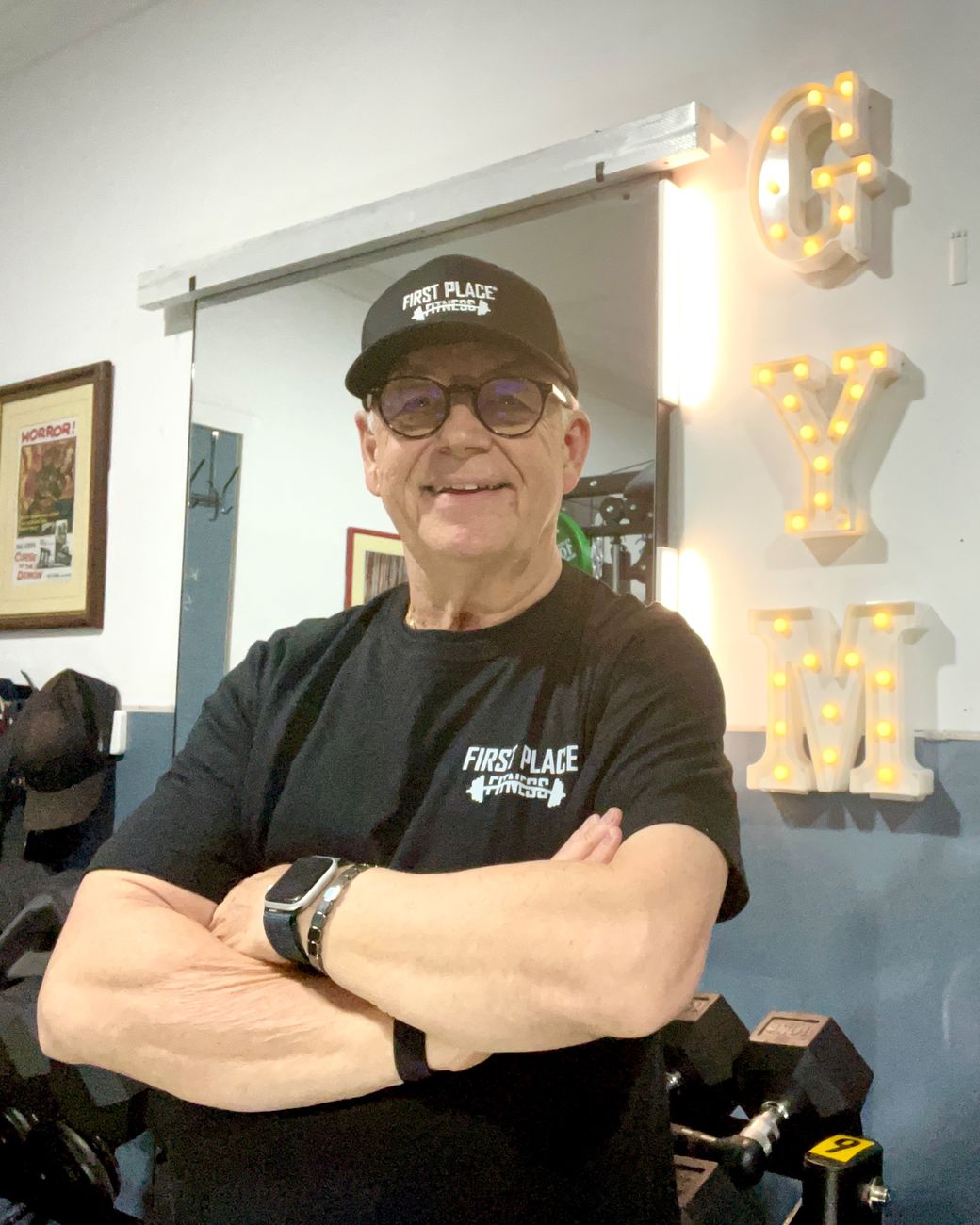 Kevin, arms folded and smiling, in his First Place Fitness cap and shirt beside the lit GYM sign in his studio