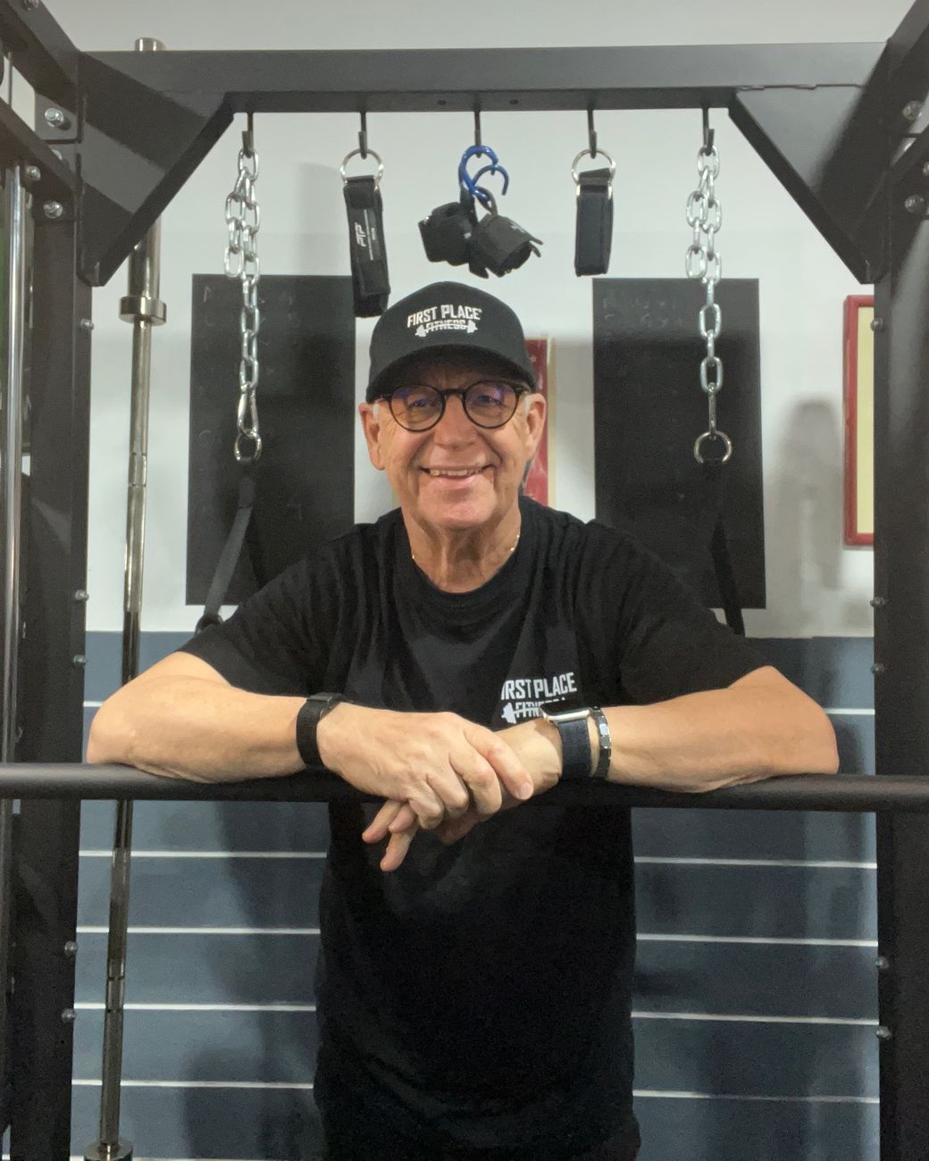 Kevin, smiling in his First Place Fitness cap and shirt, leaning on the bar of a power rack in his private studio